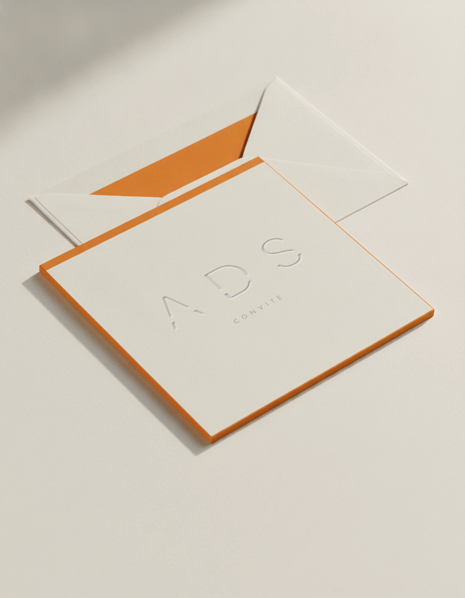 Convite do ADS em papel algodão marfim, com o logotipo em baixo-relevo e bordas pintadas em laranja, sobre um envelope com forro laranja.